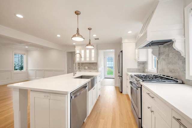 kitchen with a sink, white cabinets, appliances with stainless steel finishes, custom exhaust hood, and a center island with sink