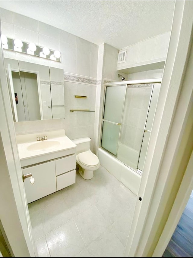 bathroom featuring a textured ceiling, toilet, bath / shower combo with glass door, vanity, and tile walls