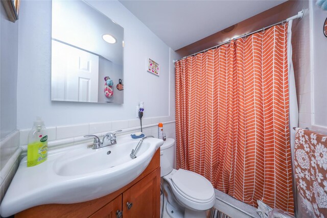 bathroom with toilet, a shower with shower curtain, vanity, wainscoting, and tile walls