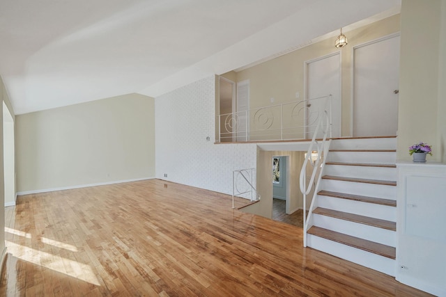 unfurnished living room featuring wallpapered walls, baseboards, lofted ceiling, wood-type flooring, and stairway