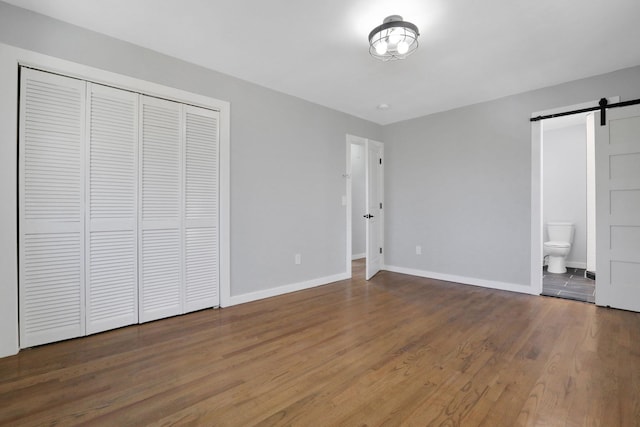 unfurnished bedroom featuring ensuite bathroom, wood finished floors, a barn door, a closet, and baseboards