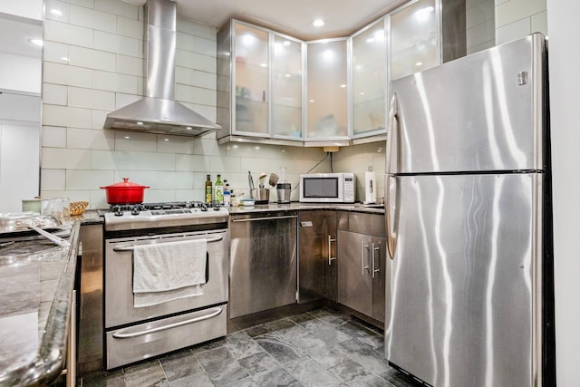 kitchen featuring stainless steel appliances, light stone countertops, wall chimney exhaust hood, tasteful backsplash, and glass insert cabinets