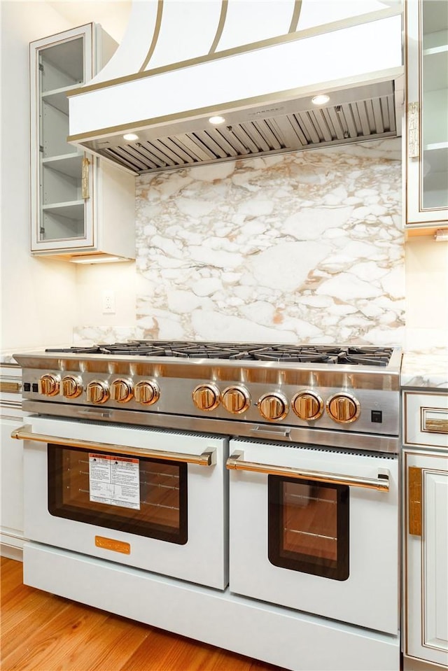 kitchen featuring light wood finished floors, tasteful backsplash, glass insert cabinets, double oven range, and exhaust hood