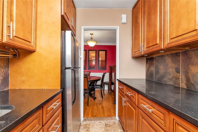 kitchen with dark stone countertops, brown cabinetry, freestanding refrigerator, ornamental molding, and decorative backsplash