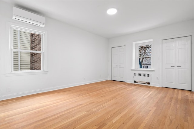 unfurnished bedroom featuring two closets, radiator, a wall mounted AC, light wood-style floors, and baseboards