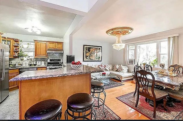 kitchen with tasteful backsplash, brown cabinetry, appliances with stainless steel finishes, a kitchen breakfast bar, and under cabinet range hood