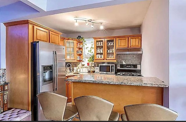 kitchen with light stone counters, under cabinet range hood, a peninsula, appliances with stainless steel finishes, and tasteful backsplash