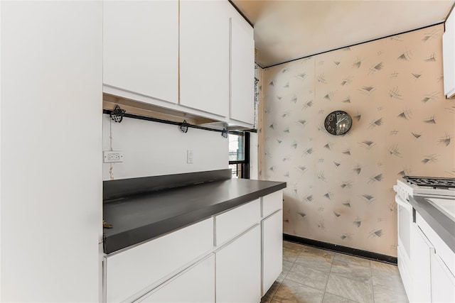 kitchen with white range with gas stovetop, baseboards, white cabinets, dark countertops, and wallpapered walls