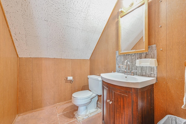 bathroom with tile patterned flooring, wooden walls, toilet, vaulted ceiling, and vanity