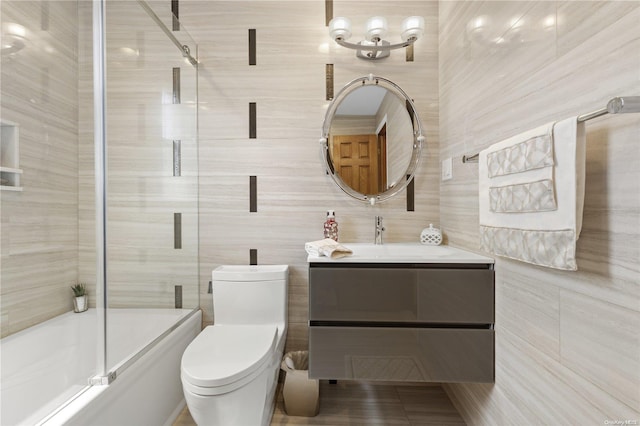 bathroom featuring bath / shower combo with glass door, vanity, toilet, and tile walls