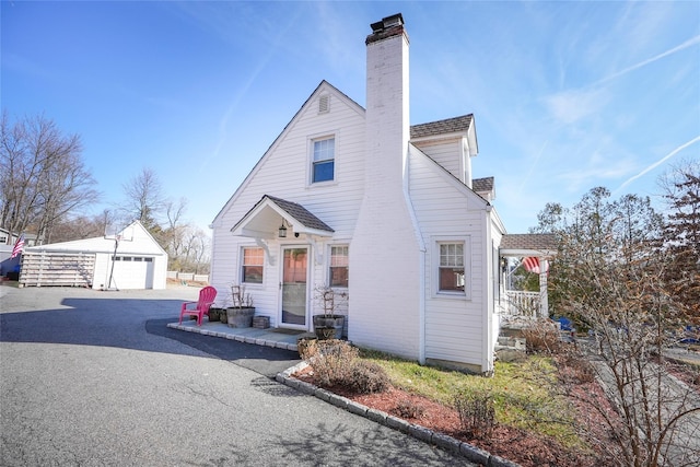 view of front facade featuring a shingled roof, a detached garage, a chimney, an outdoor structure, and driveway