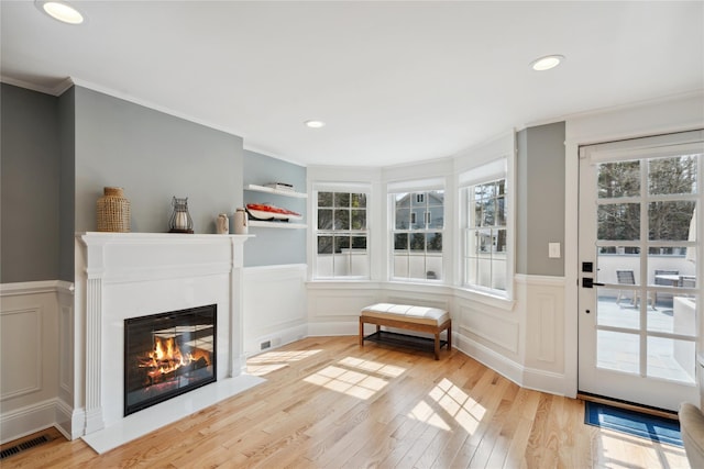 interior space featuring light wood-type flooring, a decorative wall, a glass covered fireplace, and ornamental molding