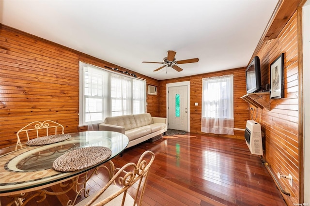 living area featuring wooden walls, dark wood-type flooring, heating unit, a wall mounted AC, and a ceiling fan