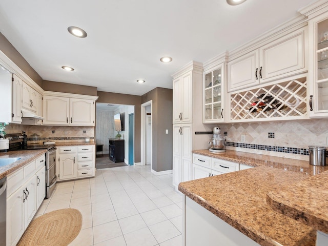 kitchen featuring light tile patterned floors, decorative backsplash, glass insert cabinets, under cabinet range hood, and appliances with stainless steel finishes