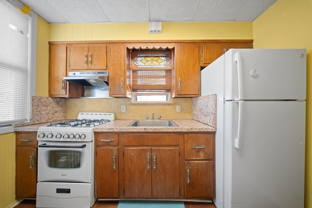 kitchen with white appliances, a sink, light countertops, under cabinet range hood, and brown cabinets