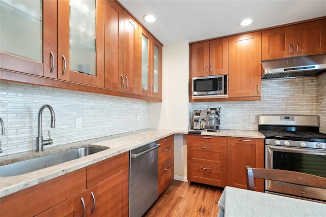 kitchen featuring light stone counters, a sink, under cabinet range hood, appliances with stainless steel finishes, and brown cabinets