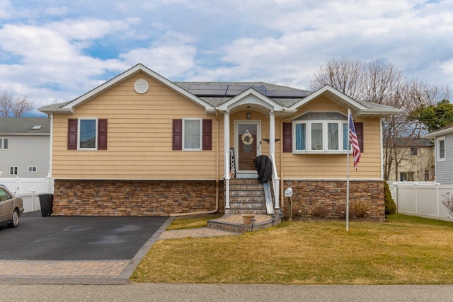 view of front of home featuring driveway, a front lawn, stone siding, fence, and solar panels