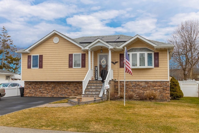 view of front facade featuring solar panels, a shingled roof, a front lawn, fence, and stone siding