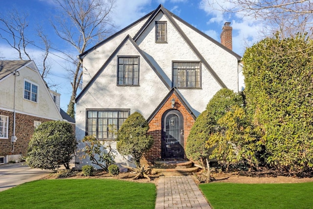 tudor-style house featuring stucco siding, a front yard, brick siding, and a chimney