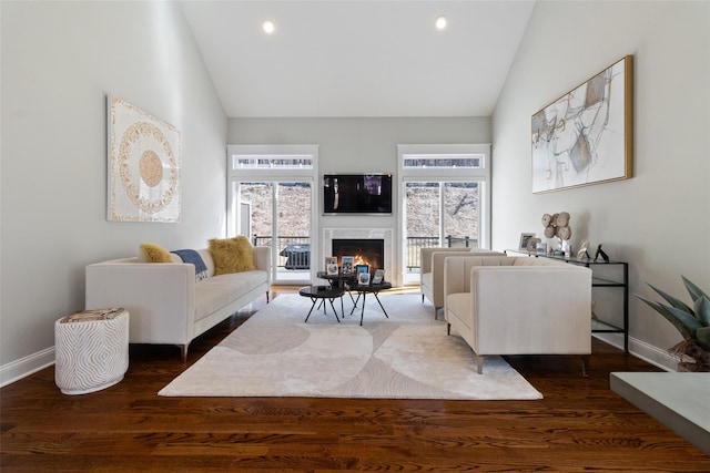 living area with dark wood-type flooring, high vaulted ceiling, a warm lit fireplace, recessed lighting, and baseboards