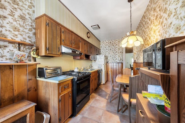 kitchen featuring wallpapered walls, under cabinet range hood, wainscoting, brown cabinetry, and black appliances