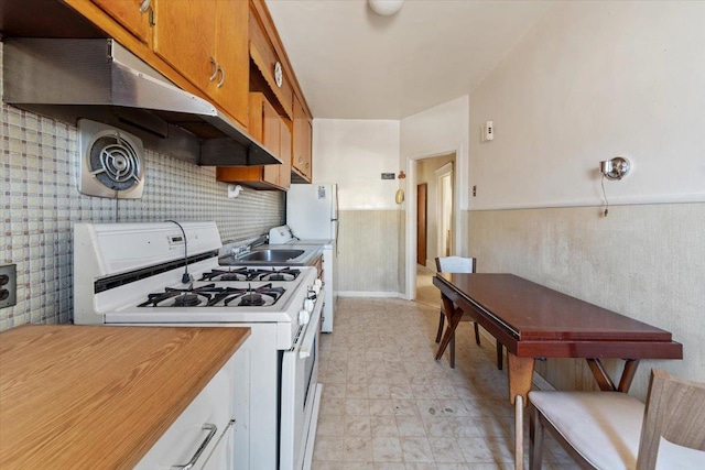 kitchen featuring under cabinet range hood, a wainscoted wall, light countertops, brown cabinetry, and white appliances