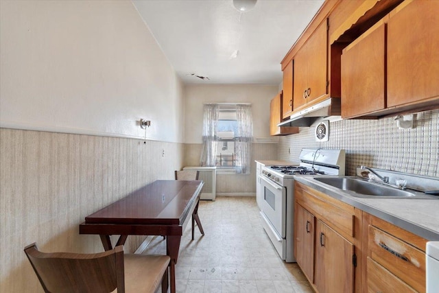 kitchen with brown cabinets, a sink, under cabinet range hood, wainscoting, and white range with gas stovetop