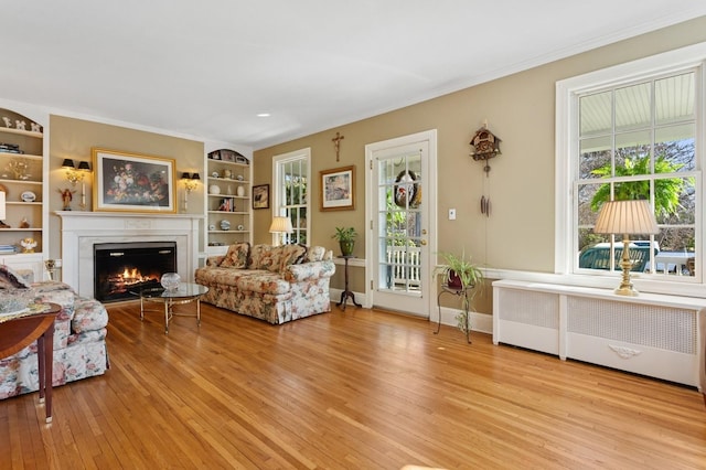 living room featuring built in shelves, radiator heating unit, light wood-style floors, a glass covered fireplace, and crown molding