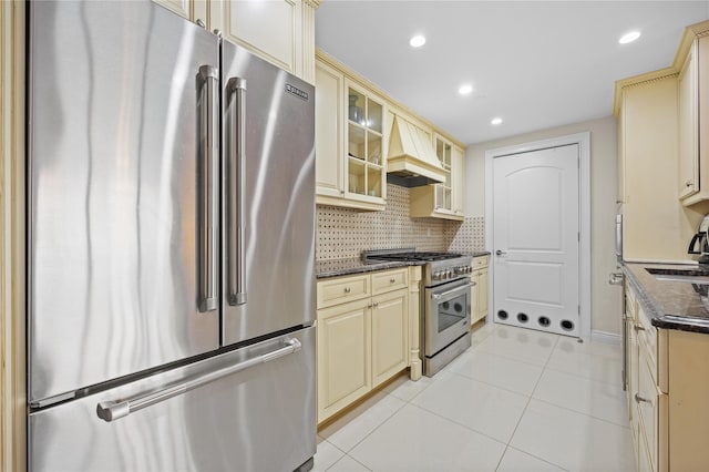 kitchen with backsplash, high quality appliances, custom range hood, light tile patterned floors, and cream cabinetry