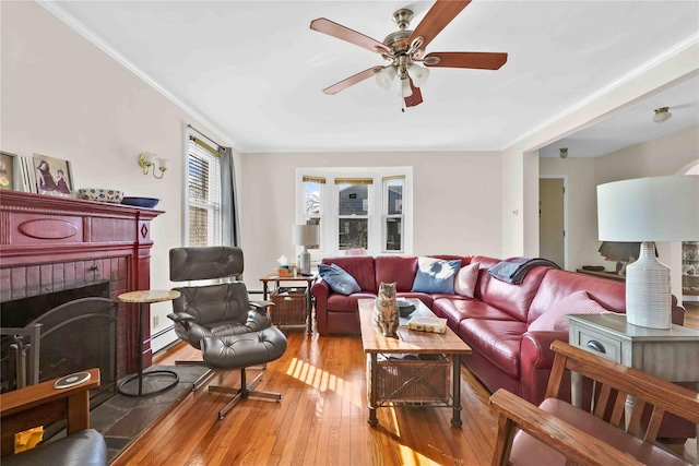 living room with a ceiling fan, hardwood / wood-style flooring, crown molding, a baseboard radiator, and a brick fireplace