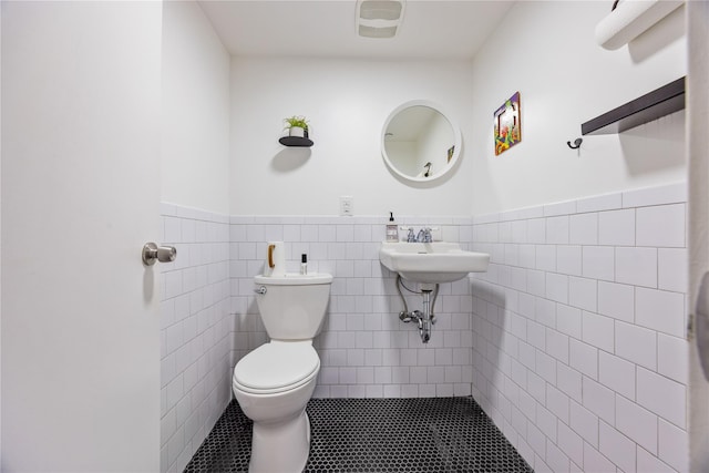 half bath with toilet, wainscoting, tile patterned floors, tile walls, and a sink