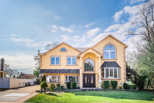 traditional home featuring stucco siding, a shingled roof, a front lawn, and fence