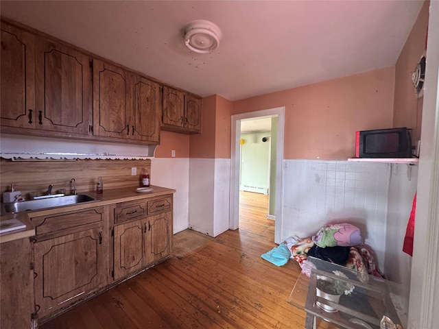 kitchen with a wainscoted wall, brown cabinets, a sink, a baseboard heating unit, and wood-type flooring