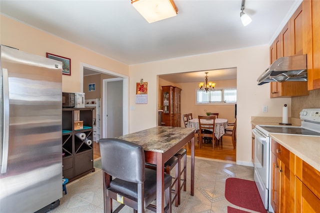 kitchen featuring brown cabinetry, freestanding refrigerator, under cabinet range hood, a notable chandelier, and white range with electric stovetop