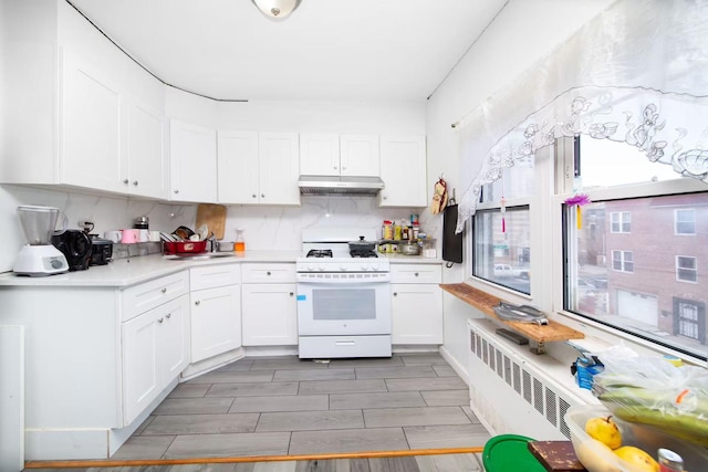 kitchen featuring under cabinet range hood, gas range gas stove, radiator heating unit, white cabinets, and light countertops