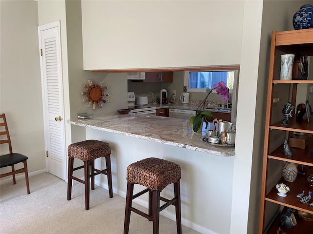 kitchen with baseboards, light stone countertops, a kitchen bar, light carpet, and white appliances