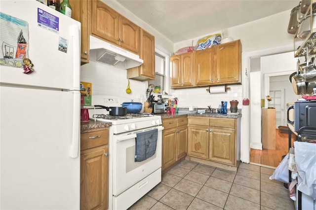 kitchen with white appliances, light tile patterned floors, under cabinet range hood, dark countertops, and backsplash