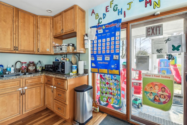 kitchen with stainless steel microwave, dark stone counters, wood finished floors, brown cabinetry, and a sink