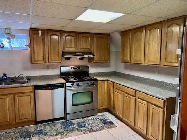 kitchen with brown cabinetry, under cabinet range hood, light tile patterned flooring, stainless steel appliances, and a sink