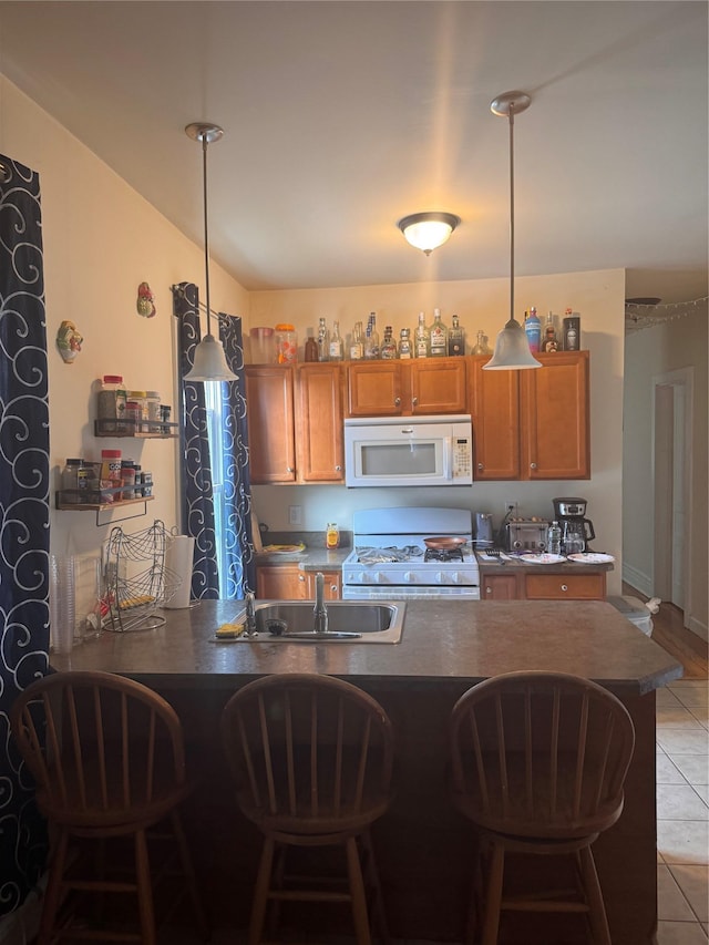 kitchen with brown cabinets, a sink, a kitchen breakfast bar, dark countertops, and white appliances