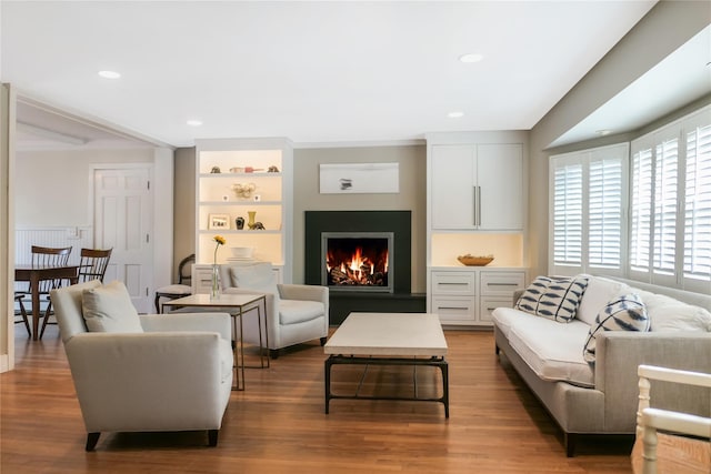 living area featuring built in shelves, recessed lighting, wood finished floors, and a warm lit fireplace