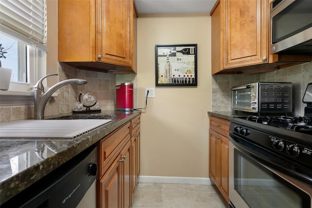 kitchen featuring brown cabinets, a sink, dark stone countertops, appliances with stainless steel finishes, and baseboards