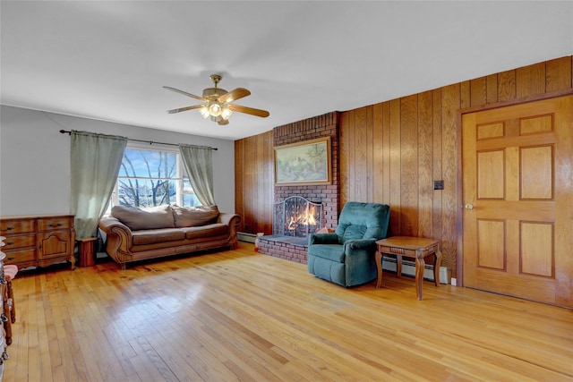 living area with a ceiling fan, light wood finished floors, a baseboard radiator, wood walls, and a brick fireplace