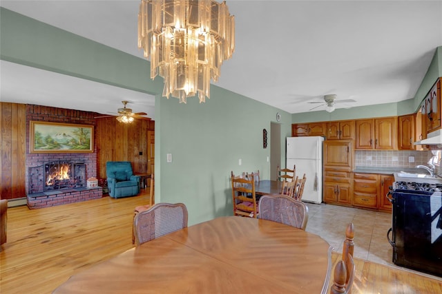 dining area featuring wooden walls, a brick fireplace, a ceiling fan, and light wood finished floors