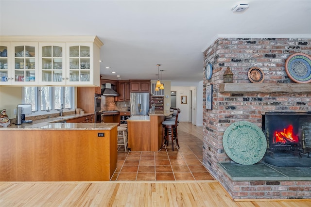 kitchen featuring glass insert cabinets, wall chimney range hood, a fireplace, stainless steel fridge, and a sink