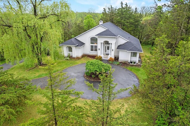 ranch-style house featuring a chimney, asphalt driveway, roof with shingles, a view of trees, and stucco siding