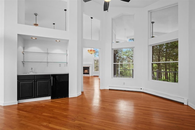 unfurnished living room with wood finished floors, wet bar, a warm lit fireplace, a baseboard radiator, and a ceiling fan