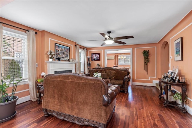 living area featuring baseboards, dark wood finished floors, a fireplace, and ceiling fan with notable chandelier