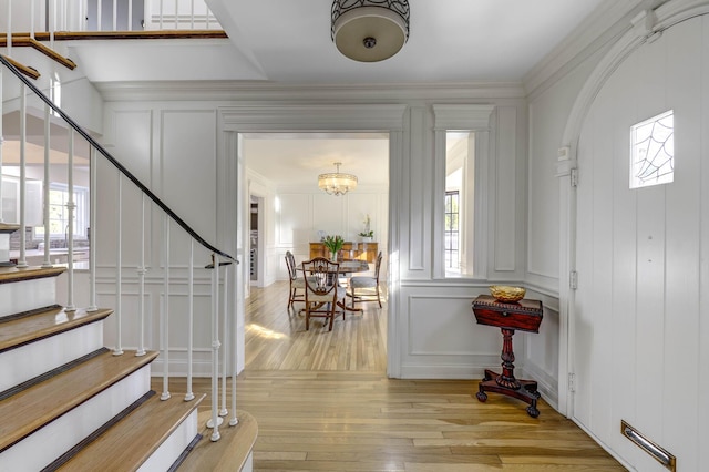 foyer entrance with light wood-type flooring, an inviting chandelier, crown molding, a decorative wall, and stairs