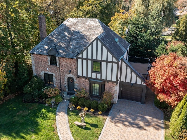 english style home with brick siding, a front yard, a chimney, decorative driveway, and a garage